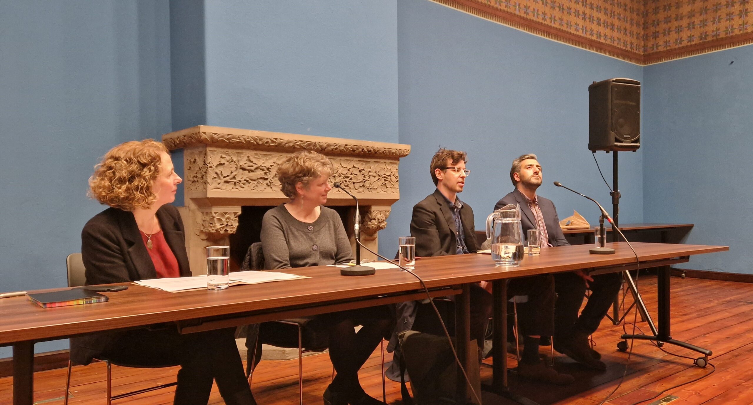 Two women and two men are sitting at a table in front of microphones while one is talking.